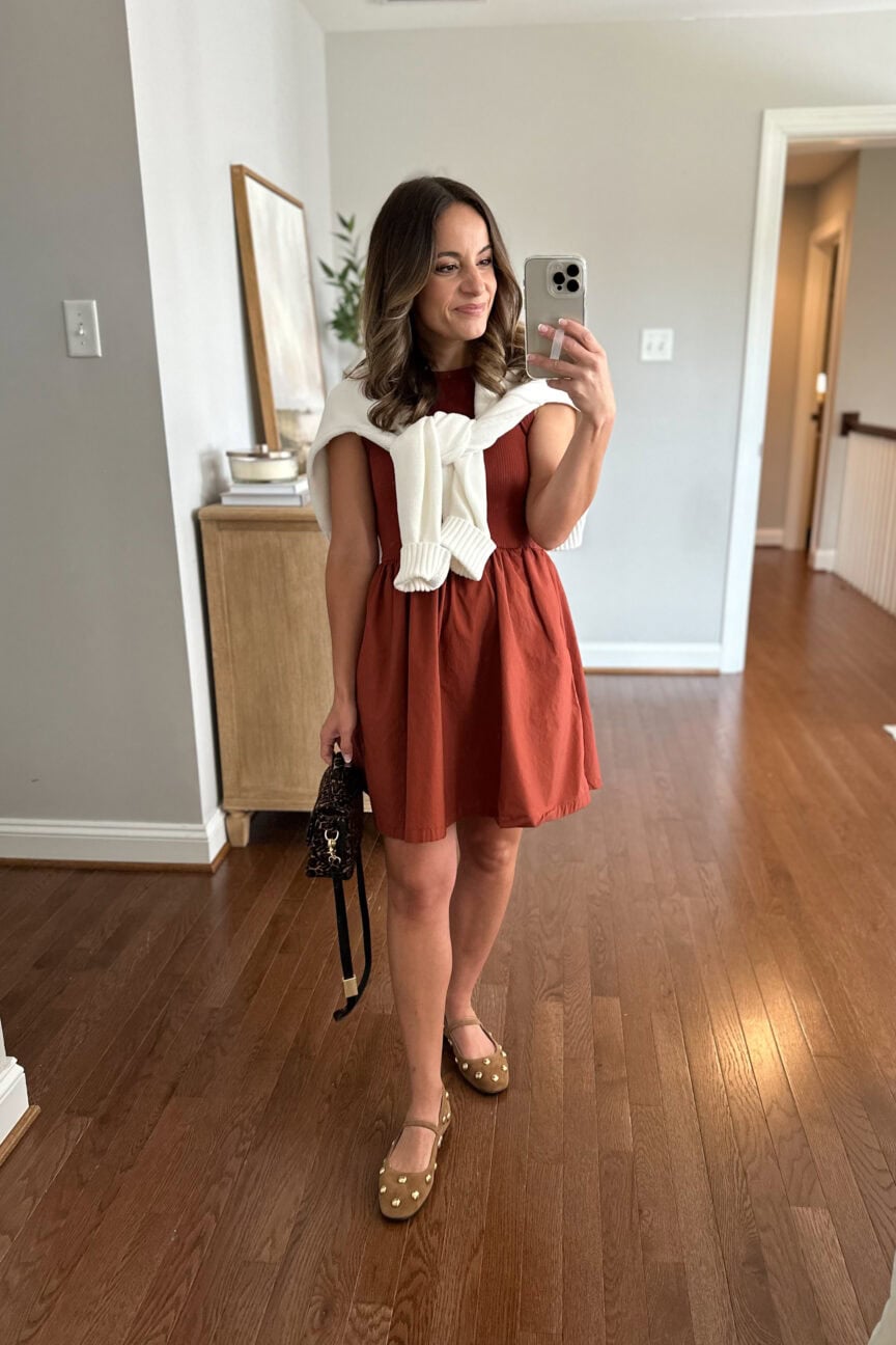 Early fall outfit featuring a red dress styled with a white sweater draped over the shoulders and studded ballet flats, showcasing a chic transitional look and fall shoe trend